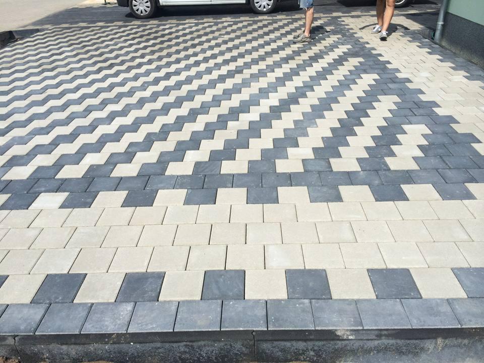 Ułożona nawierzchnia z kostki brukowej w geometryczny wzór szewronu, wykorzystująca szare i jasnoszare kostki, z widocznymi fragmentami samochodu i dwóch osób w tle.