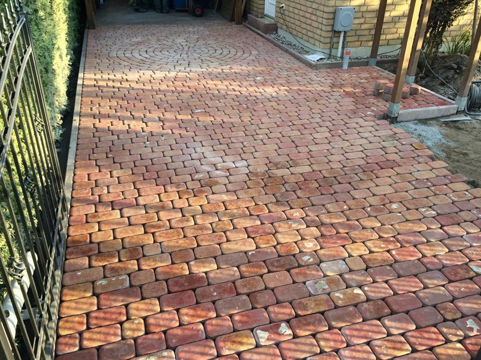 Czerwono-brązowa kostka brukowa ułożona na podjeździe z okrągłym wzorem w tle, widoczne elementy ogrodzenia i fragment konstrukcji z drewna.