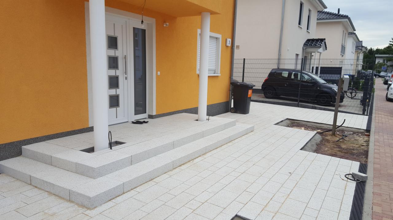 Wykończone schody wejściowe i częściowo ułożony chodnik z jasnoszarej kostki brukowej na posesji z żółtą elewacją domu, widoczny fragment ogrodzenia.