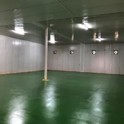 Pusta, oświetlona hala magazynowa z zieloną podłogą, białymi ścianami z blachy falistej i wentylatorami w ścianie.