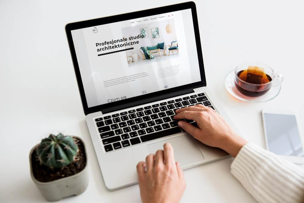 Osoba pisze na laptopie prezentującym stronę internetową 'Profesjonalne studio architektoniczne', obok filiżanka herbaty i smartfon, w tle białe biurko i kaktus w betonowej doniczce.