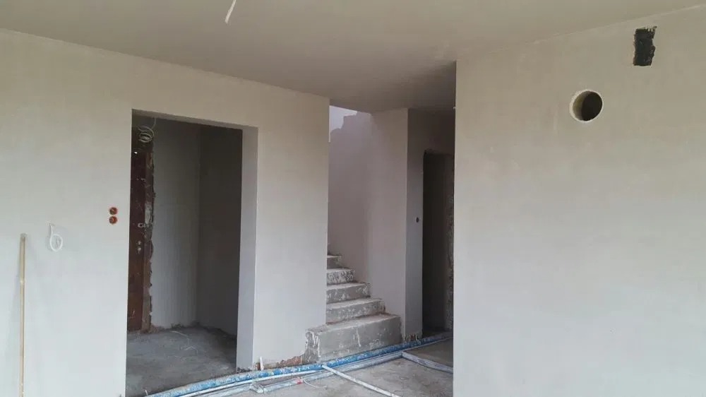 Wnętrze domu w trakcie wykańczania, ściany po szpachlowaniu, widoczne otwory drzwiowe, schody w tle, niebieskie rury na podłodze.