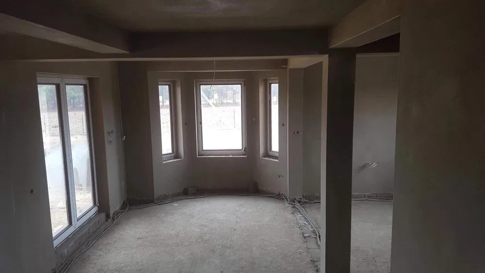 Surowy stan wnętrza z oknami przed pracami wykończeniowymi, widoczne kable i ściany przygotowane pod dalsze prace.