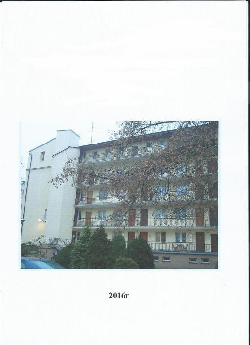 Wielopiętrowy budynek mieszkalny z balkonami, widoczny z poziomu gruntu, częściowo zasłonięty przez drzewa i krzewy.