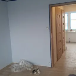 Świeżo pomalowane na jasnoniebiesko ściany z niebieską taśmą malarską przy suficie i ościeżnicach, widoczne narzędzia malarskie i wiaderka z farbą na podłodze z paneli imitujących drewno.