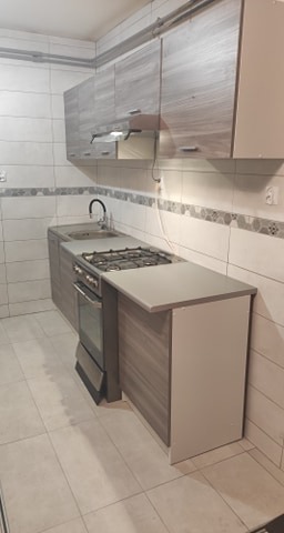 Nowoczesna kuchnia z szarymi szafkami, jasnymi płytkami i wbudowaną kuchenką gazową z piekarnikiem, widok z boku na fragment blatu roboczego.