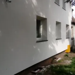 Boczny widok budynku po wykonaniu elewacji, z widocznymi oknami, fragmentem rusztowania po lewej stronie i drzewem po prawej, z żółtą skrzynką gazową przy ścianie.