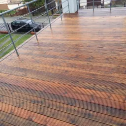 Mokry, drewniany taras z metalową barierką, widok na budynki mieszkalne i zaparkowany samochód w tle.