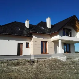 Nowo wybudowany dom jednorodzinny z kremową elewacją, czarnym dachem i balkonem, widoczny w stanie surowym otoczenia.