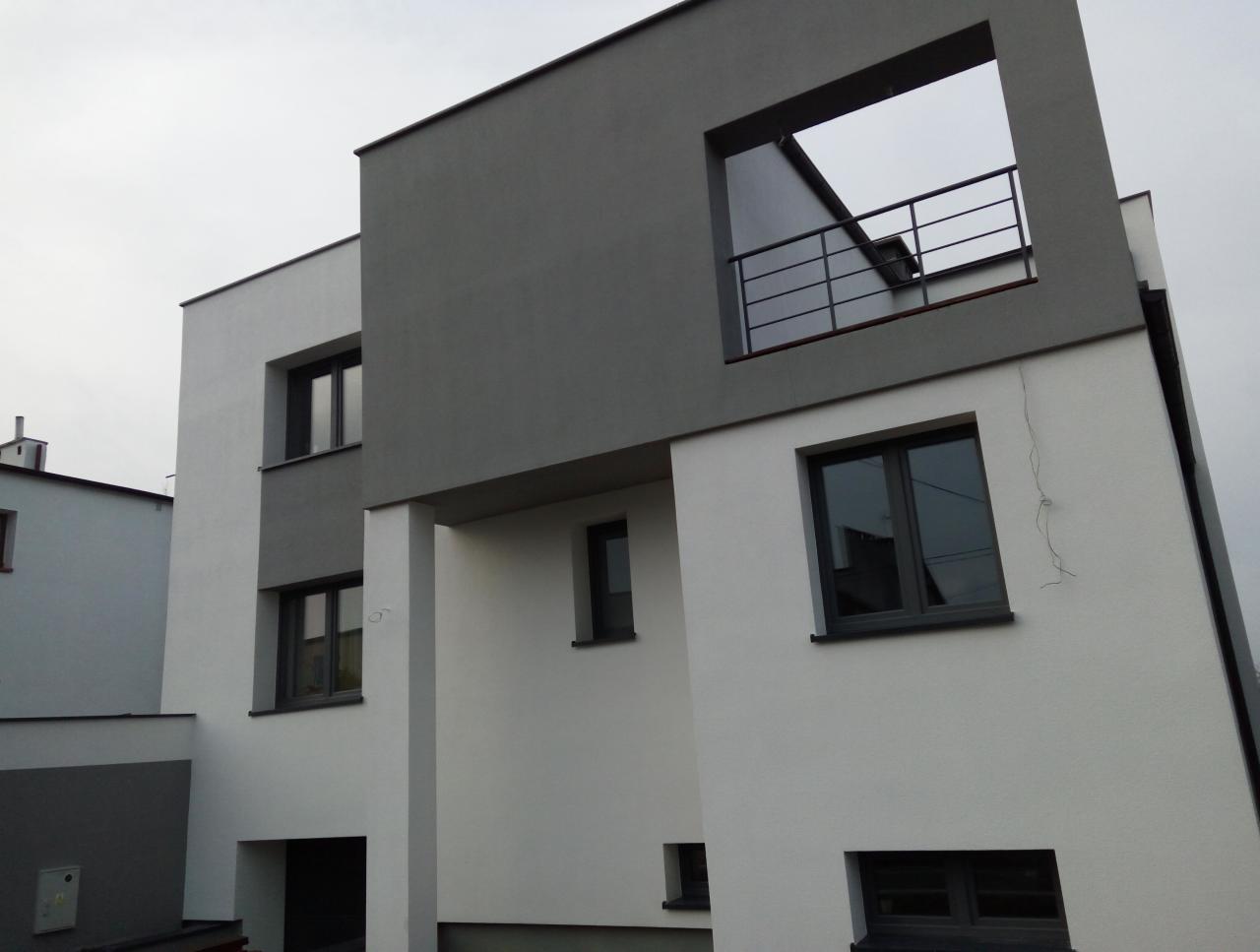 Nowoczesny dom z elewacją w odcieniach szarości i bieli, z minimalistycznym balkonem i wyraźnymi geometrycznymi kształtami.