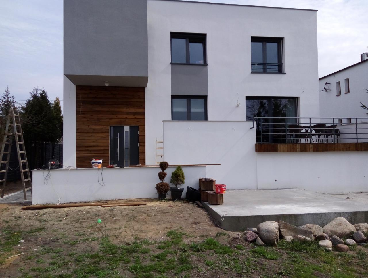 Nowoczesny dom z częściowo wykończoną elewacją: biały tynk, szara wstawka i drewniana okładzina przy wejściu. Widoczne drabiny, puszki z farbą i elementy dekoracyjne.