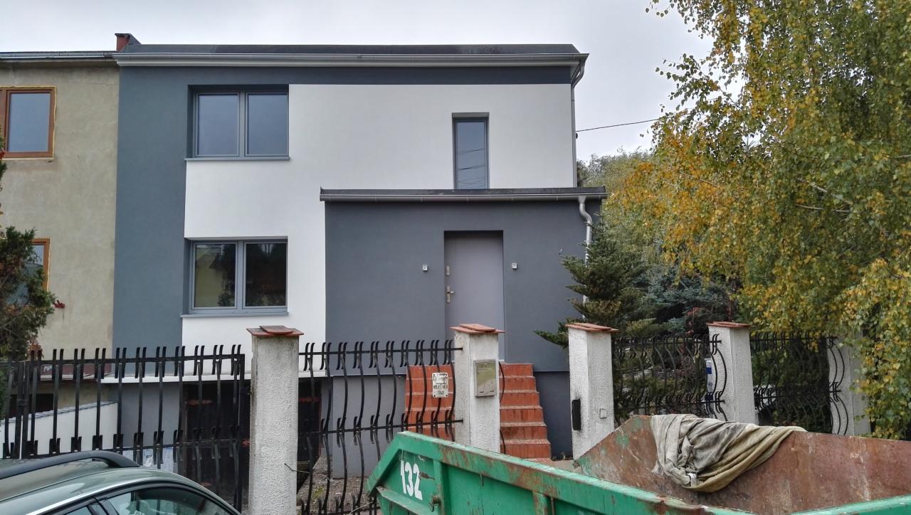 Dwukondygnacyjny dom z elewacją w odcieniach szarości i bieli, widok z zewnątrz, z zielonym kontenerem na gruz z przodu.