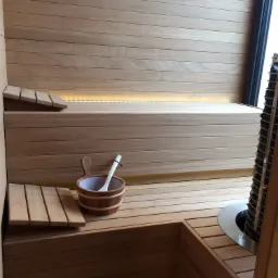 Wnętrze wykończonej drewnem sauny z jasnymi deskami, kubełek z chochlą na dolnej ławce, grzejnik z kamieniami w rogu.