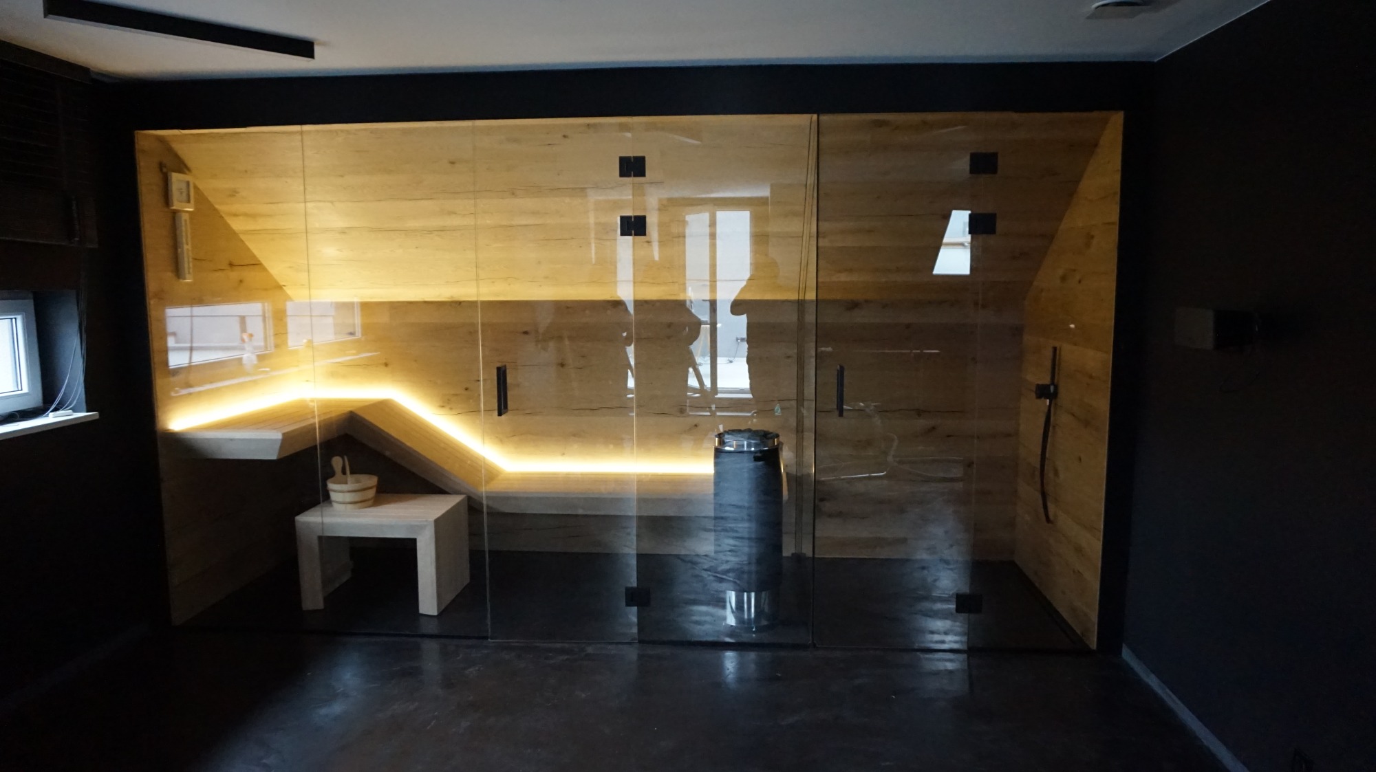 Nowoczesna kabina z drewna i szkła z podświetleniem LED, wiaderkiem i czerpakiem na półce, oraz czarnym, metalowym piecem.