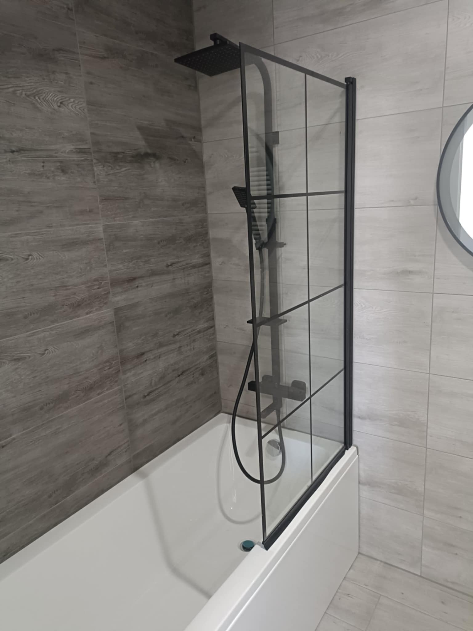 Nowoczesna kabina prysznicowa z czarnymi profilami na wannie, szare płytki imitujące drewno na ścianie. Minimalistyczny design łazienki.