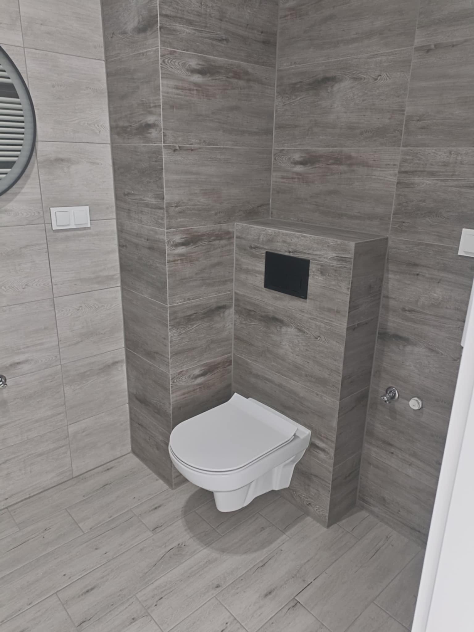 Nowoczesna łazienka z szarą, drewnopodobną glazurą. Podwieszana toaleta z zabudową. Minimalistyczny styl, czyste linie, eleganckie wykończenie.