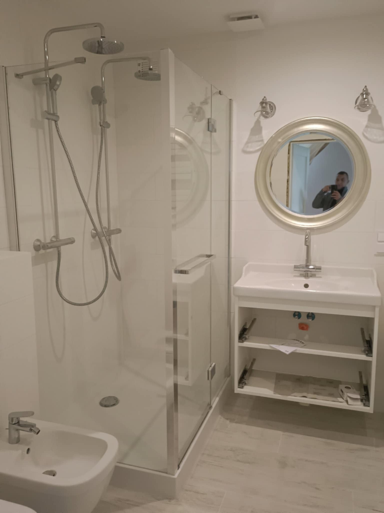 Biała łazienka po remoncie: kabina prysznicowa ze szklaną ścianą, umywalka z szafką i okrągłe lustro. W lustrze odbija się osoba robiąca zdjęcie.