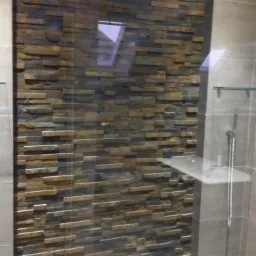 Nowoczesna kabina prysznicowa z wykończeniem z kamienia dekoracyjnego na jednej ze ścian, szklane półki i chromowana armatura.