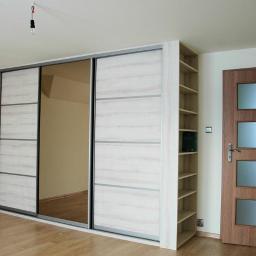 Clone-Meble - Wnętrze pokoju z dużą szafą przesuwną z lustrem, regałem z półkami i drewnianymi drzwiami z przeszkleniami, widok na podłogę z paneli.