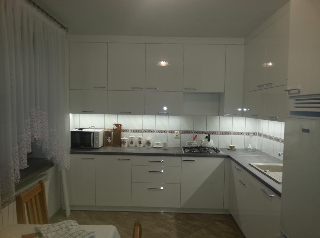 Jasna kuchnia z białymi, błyszczącymi frontami szafek, ciemnym blatem i dekoracyjnym pasem płytek nad blatem, widoczne akcesoria kuchenne i zasłona w oknie.