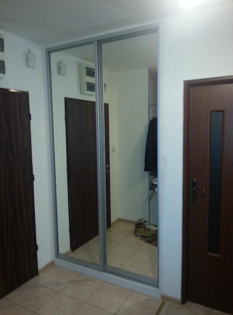 Przesuwne drzwi szafy z lustrami w przedpokoju, widoczne odbicie sąsiednich drzwi i fragment szafy wnękowej, jasne płytki podłogowe.