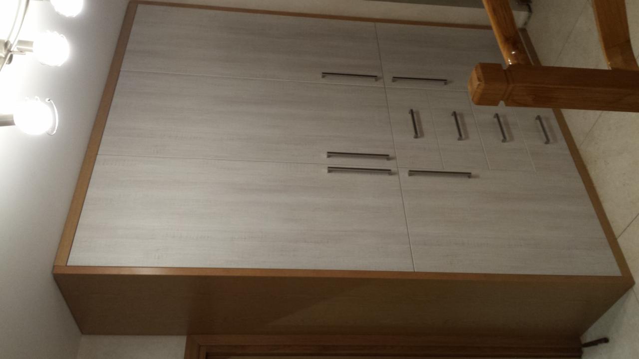 Szafka łazienkowa z frontami w kolorze jasnego drewna, z podłużnymi metalowymi uchwytami, oświetlona przez jasne światło lampy, widoczna z dołu pod kątem.