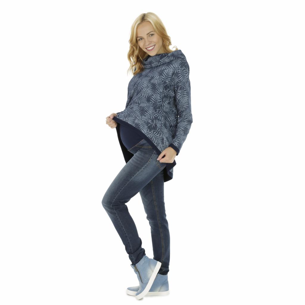 Uśmiechnięta blondynka w ciążowej bluzie z wysokim golfem w liście, dżinsach i sportowych butach na koturnie, pozuje na białym tle.