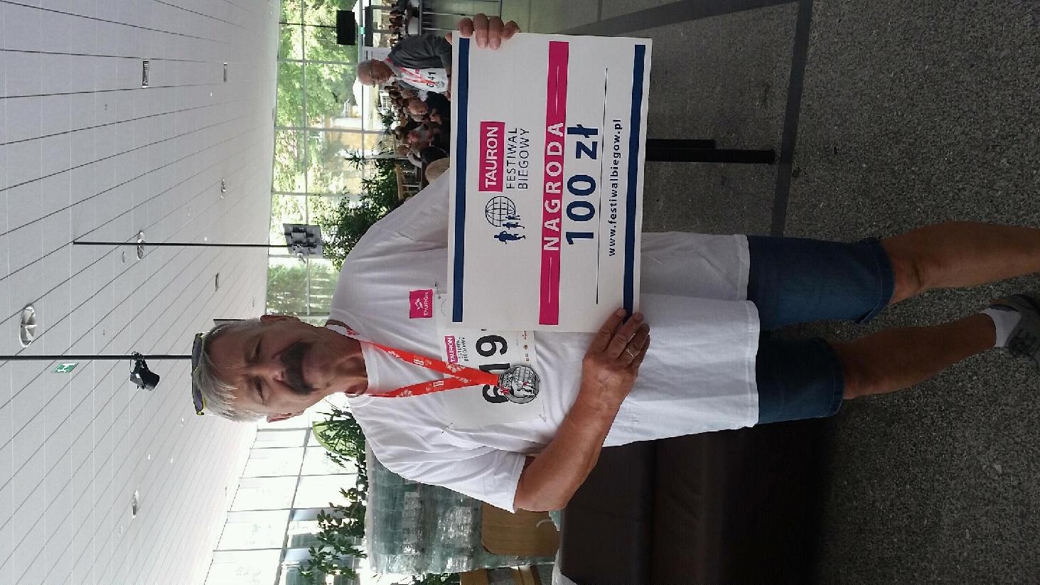 Mężczyzna z medalem i tablicą 'Nagroda 100 zł' na tle wnętrza hali podczas Tauron Festiwalu Biegowego. Uśmiechnięty, w białej koszulce i dżinsowych spodenkach.