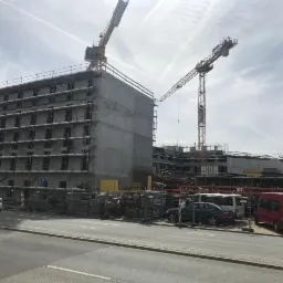 Wznoszenie surowego budynku wielokondygnacyjnego z betonu z widocznymi rusztowaniami i dwoma żurawiami wieżowymi na tle pochmurnego nieba.