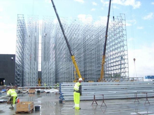 Montaż szkieletu hali magazynowej z użyciem dwóch dźwigów, widoczni pracownicy w odblaskowych ubraniach, na ziemi leżą profile stalowe.