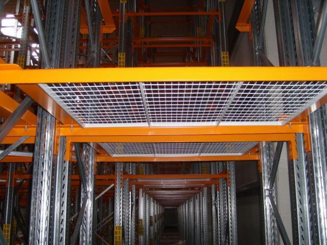 Wnętrze magazynu z pomarańczowymi regałami i metalowymi kratami tworzącymi poziome powierzchnie, widok w głąb korytarza między regałami.