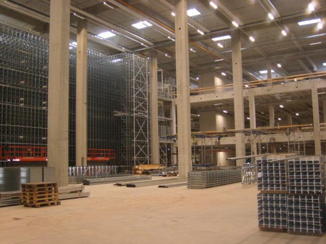 Wnętrze hali w trakcie budowy z widocznymi betonowymi kolumnami, stalową konstrukcją regałów magazynowych i ułożonymi profilami stalowymi na podłodze.