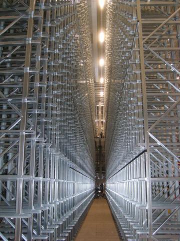 Wnętrze wysokiego magazynu z metalowymi regałami, widok w perspektywie wzdłuż korytarza, oświetlenie sufitowe.