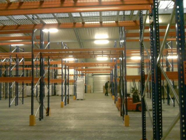 Wnętrze hali magazynowej z regałami paletowymi w trakcie montażu, widoczny wózek widłowy i pracownicy w oddali.