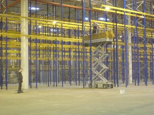 Montaż regałów magazynowych: pracownicy na podnośniku koszowym instalują żółte belki w niebieskich słupkach w przestronnej hali magazynowej, malarz zabezpiecza filar