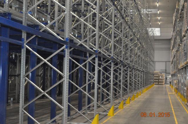 Wnętrze magazynu z wysokimi regałami stalowymi, zabezpieczonymi żółtymi odbojnikami, widok wzdłuż korytarza z paletami w tle.