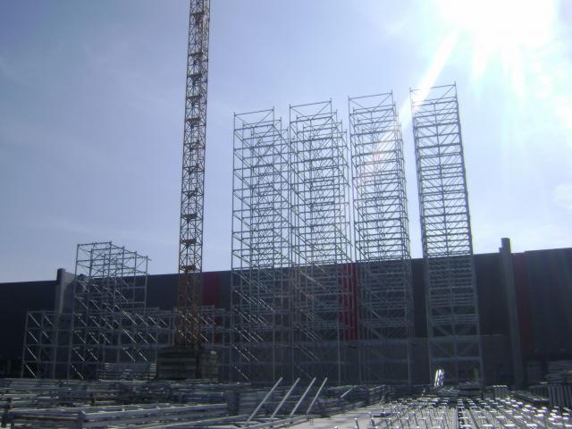 Montaż szkieletu hali stalowej z użyciem dźwigu, widoczne elementy konstrukcyjne na placu budowy, błękitne niebo w tle.