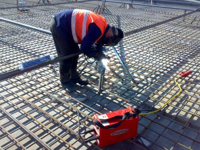 Spawacz w pomarańczowej kamizelce spawa stalową siatkę zbrojeniową na budowie, widoczna spawarka Fronius i opakowanie prętów zbrojeniowych.