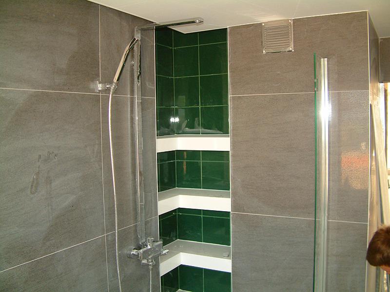 Nowoczesna łazienka z szarymi płytkami imitującymi kamień, zabudowana wnęka z zielonymi płytkami i białymi półkami, chromowana bateria prysznicowa z deszczownicą.