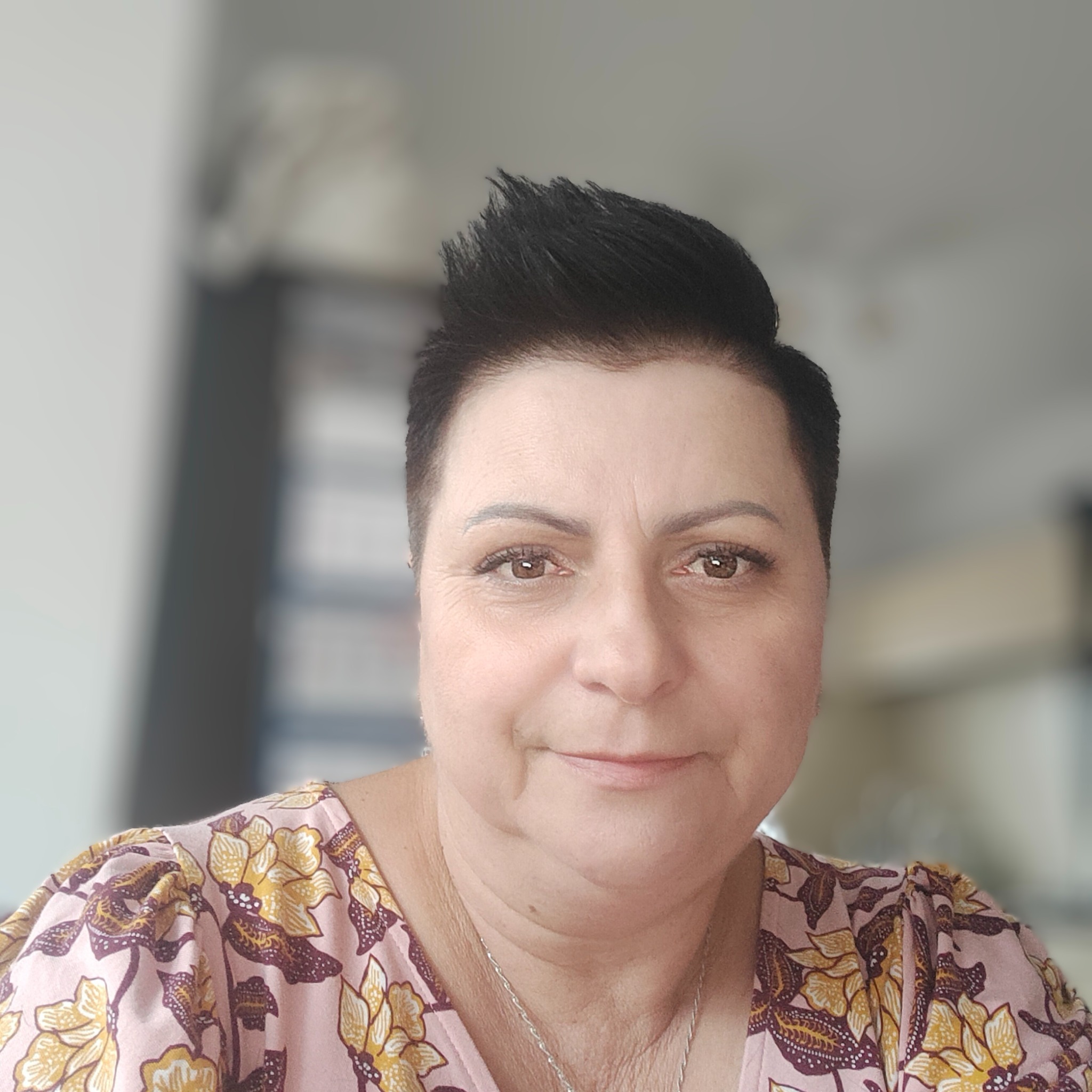 Portret kobiety z krótkimi, ciemnymi włosami, ubranej w bluzkę w kwiaty, z delikatnym uśmiechem i srebrnym łańcuszkiem na szyi, w tle rozmyte wnętrze.