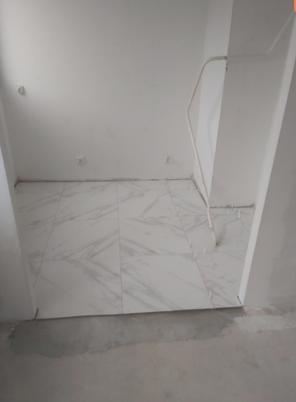 Białe marmurowe płytki podłogowe ułożone w pomieszczeniu, z widocznymi krzyżykami dystansowymi i fragmentem rury przy ścianie, surowy beton na podłodze przed wejściem do pomieszczenia.