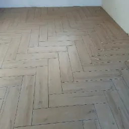 Ułożona w jodełkę jasna, podłużna terakota imitująca drewno, krzyżyki dystansowe widoczne w spoinach, zabezpieczona folią ściana w tle.