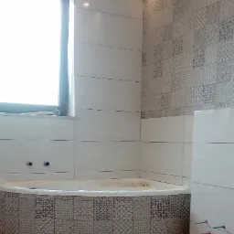 Niedokończona łazienka z wanną obudowaną mozaiką, białe płytki na ścianach i wzorzysta ściana w tle, widoczne wystające rury instalacyjne.