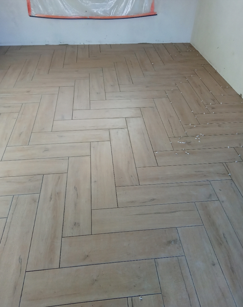 Ułożona w jodełkę jasna, podłużna terakota imitująca drewno, krzyżyki dystansowe widoczne w spoinach, zabezpieczona folią ściana w tle.