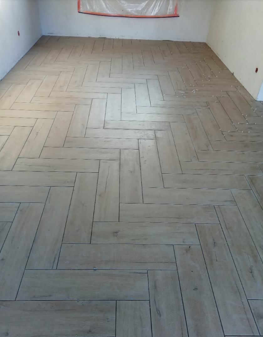 Układanie podłogi z płytek imitujących drewno w jodełkę, widoczne fugi i częściowo zabezpieczona folią ściana.