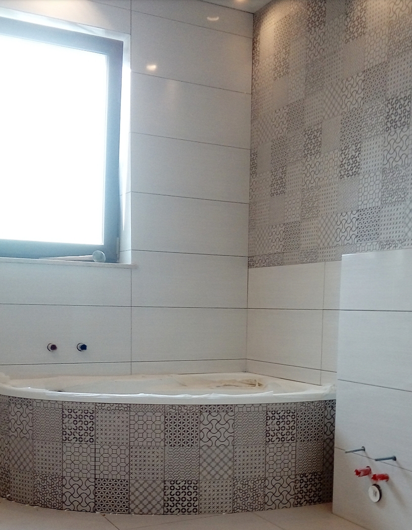 Niedokończona łazienka z wanną obudowaną mozaiką, białe płytki na ścianach i wzorzysta ściana w tle, widoczne wystające rury instalacyjne.