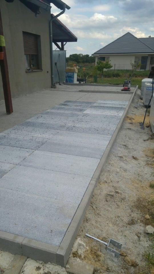 Układanie kostki brukowej na posesji, widoczna ubijarka, paleta z kostką i narzędzia, w tle dom jednorodzinny.