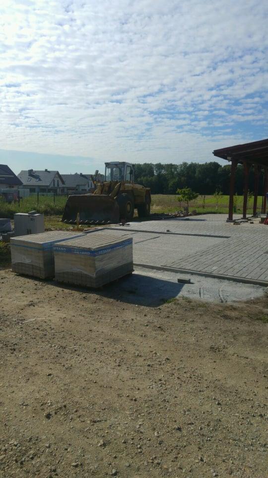 Żółta ładowarka kołowa stoi na placu budowy, obok ułożona kostka brukowa i palety z nową kostką, w tle domy i drzewa.