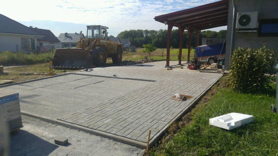 Żółta ładowarka kołowa stoi na placu budowy, gdzie układana jest kostka brukowa. Część powierzchni jest już wyłożona, a pozostała część jest przygotowana pod kostkę. W tle widoczny dom i zadaszona...