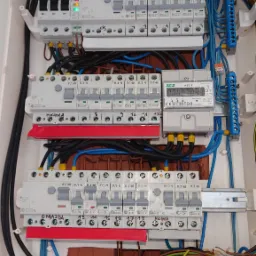 Otwarta skrzynka rozdzielcza z bezpiecznikami, licznikiem energii i licznymi przewodami w różnych kolorach, widoczne oznaczenia obwodów 'Kuchnia' i 'Klima' na naklejkach.